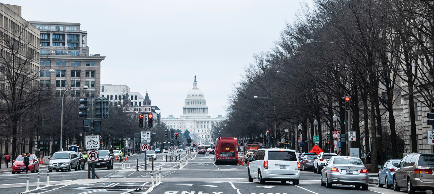 Daytime view along busy Constitution Avenue, Washington DC. Cars and buses wait at red lights, the Capitol building providing a striking backdrop.