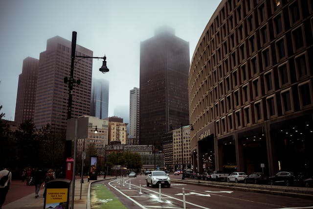 Street side image towards Boston’s CBD skyline during the daytime. People are walking on the sidewalk, cars are driving down the street, and the tops of the CBD’s skyscrapers are obscured by fog. Image at Offices.net.
