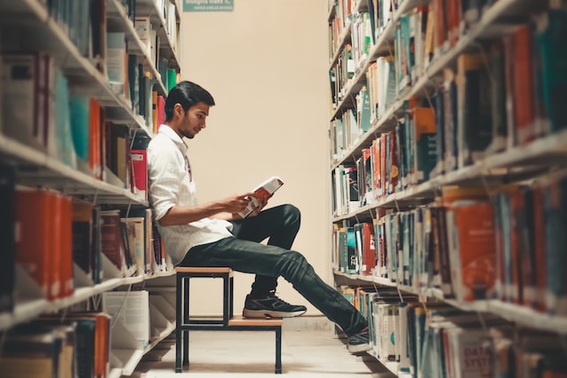 male student sitting on step stool and reading in library aisle image at offices.net