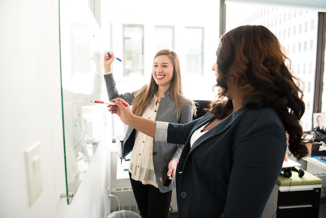 two female colleagues smiling and writing on a whiteboard image at offices.net