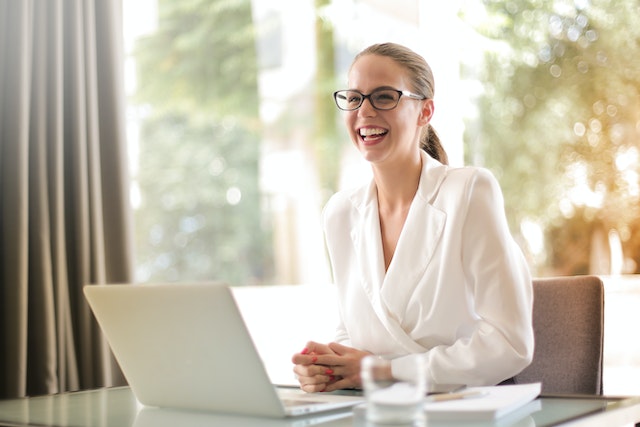 a smiling young woman wearing glasses and a white business top sitting at a desk with a white opened laptop computer image at ofices.net