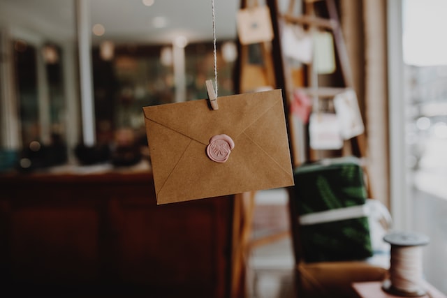 an envelope closed with a pink wax seal is suspended by a wooden clothes peg and string attached to a ceiling that is out of the shot image at offices.net