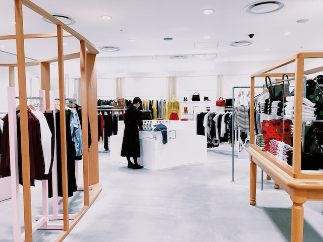 a female fashion store worker in a black dress stands at a white counter in the center of the store with clothes racks arrayed around it image at offices.net