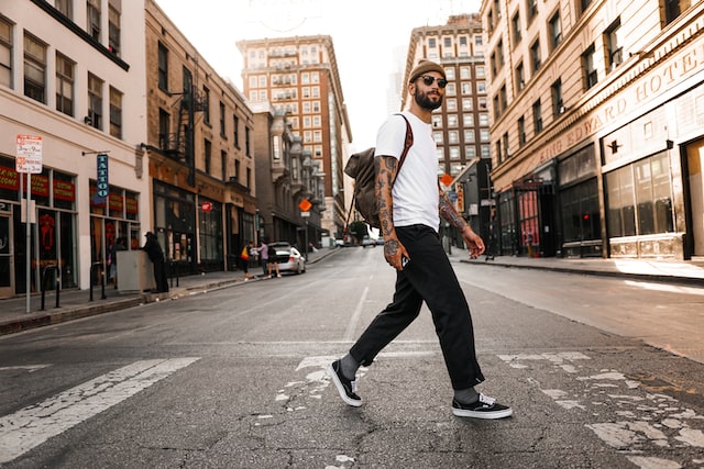 a trendy looking bearded and bespectacled man with tattoos and a backpack walking across a crosswalk on a quiet urban street with shops and hotels lining the street image at offices.net
