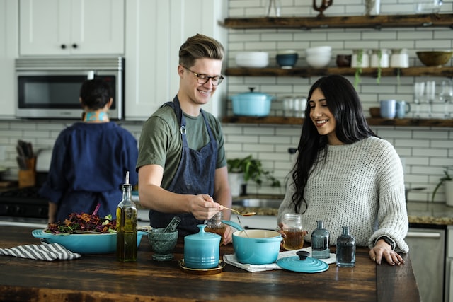 two smiling colleagues one male and one female work on a recipe together with the man measuring out a spoonful of an ingredient before adding it to a pot on the kitchen bench displaying how cooking classes can be one of the fresh and fun in-office event ideas image at offices.net