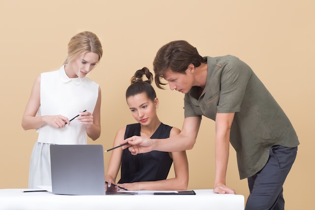 a young woman sits at a white desk with her silver colored laptop open in front of her and a male and female colleague standing to either side of her with the male colleague resting his hand on the desk and pointing a pencil at the sitting colleague's screen image at offices.net