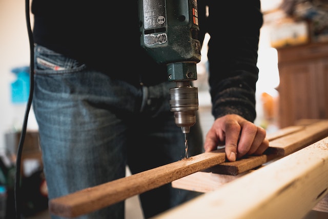 close up of drill being held by a man that is starting to drill through a long and slender plank of wood image at offices.net