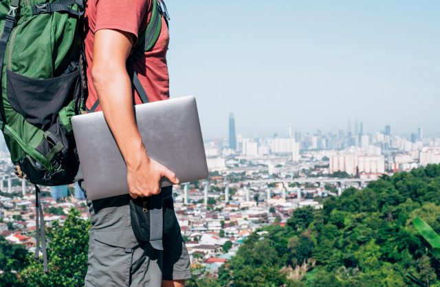 close up of a man wearing a large backpack and holding a closed silver laptop on top of a lush green hill that overlooks a large urban area that stretches out to the horizon image at offices.net