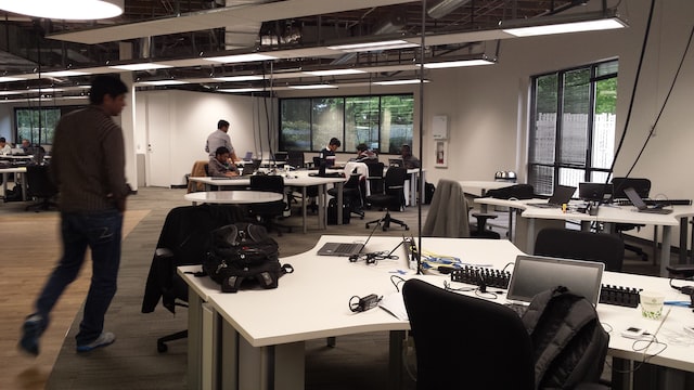 a moderately lit action scene of an office with a man walking in the foreground past a communal desk with other desks in the background with people sitting at them image at offices.net