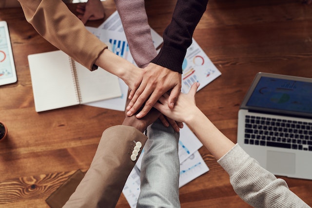 top down close up of six people arranged in a circle around a desk placing their hands on top of each other above the desk's center displaying agreement at fresh and fun in-office event ideas image at offices.net