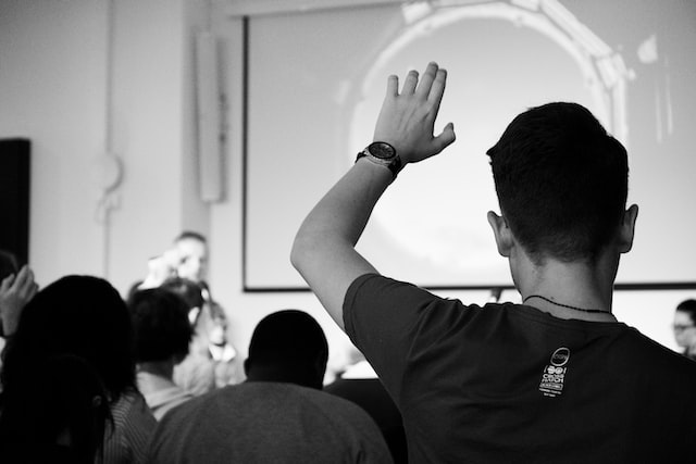 a dark haired man in a presentation audience has his hand raised at audience question time with a panel of speakers sitting on the stage in front of him image at offices.net