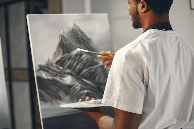 a standing dark hired man in a white tee shirt holding a palette in one hand and a brush in the other painting a black and white mountainous scene image at offices.net