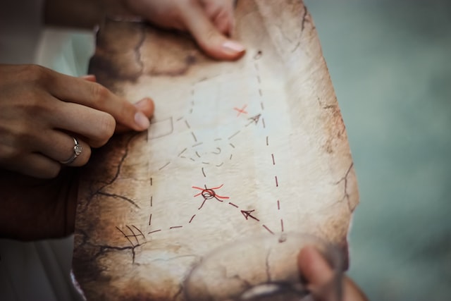 two hands holding a weathered looking treasure map with two hands pointing at a square shaped location on the center of the map demonstrating a treasure hunt as one of the fresh and fun in-office event ideas image at offices.net