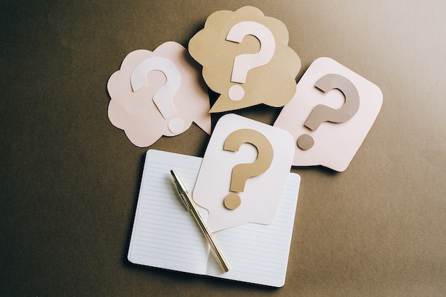 an open blank notepad with a gold colored pen resting on it lays on a brown desk with paper cut outs of speech bubbles with question marks in them around the notebook image at offices.net