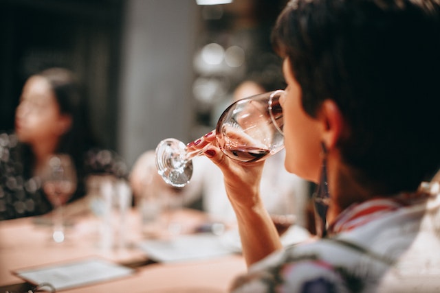 a close up from behind the shoulder of a seated woman with short dark hair and long earrings sipping red wine out of a wineglass with other people sitting around her discussing the wine image at ofices.net