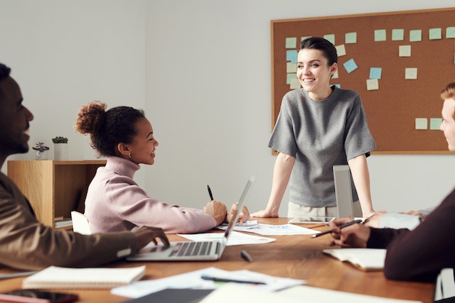 Smiling office colleagues conduct a meeting discussing United States commercial property prices per square foot. Three people are seated with their laptops open in front of them on the meeting room table while one woman in a grey top stands at the head of the table addressing her gathered colleagues - Offices.net.