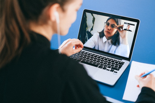 A woman sitting at a blue desk and taking notes with pen and paper is on a video call with a female colleague who is wearing a white business shirt and glasses and looks concerned. Image at Offices.net.