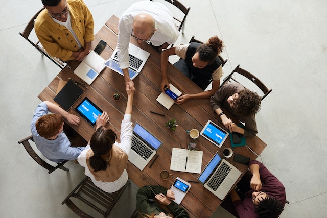 Bird's-eye view of eight colleagues sitting around all sides of a rectangular meeting desk with laptops, tablets, and note-taking implements resting in front of each. Two, a man and woman, are standing and shaking hands across the desk image at Offices.net.