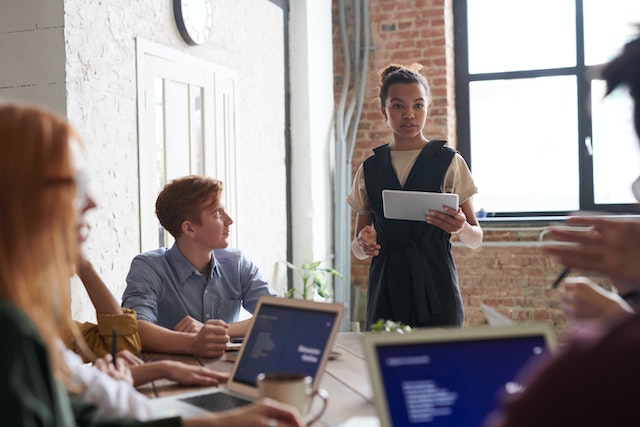 A young woman holding a tablet backlit by sunlight coming through a window confidently addresses her colleagues sitting around her at a table with laptops open in front of them or with notepads and pens for taking notes. Image at Offices.net.