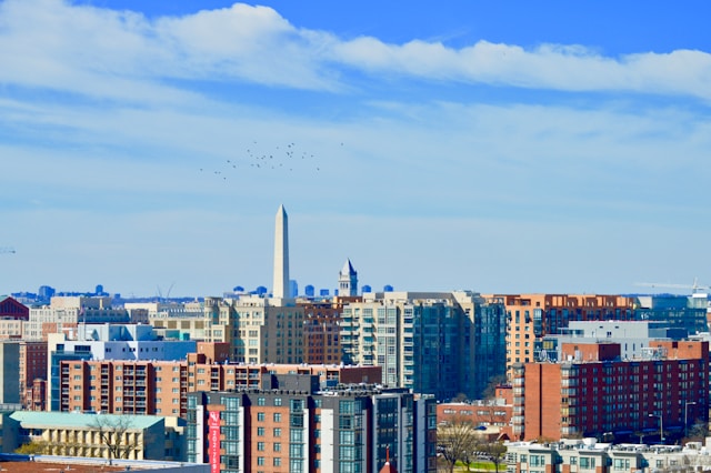 A view across the Washington DC skyline at daytime. The Washington Monument rises in the distance above many-hued buildings towards a blue sky streaked with white clouds. Image at Offices.net.