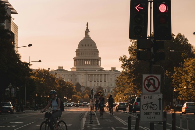 A dusk street-level view down a bustling street towards the U.S. Capitol Building, framed by a pastel orange sky. The street is busy with cars on both sides, and many cyclists ride down the middle in a cycling lane. Image at Offices.net.