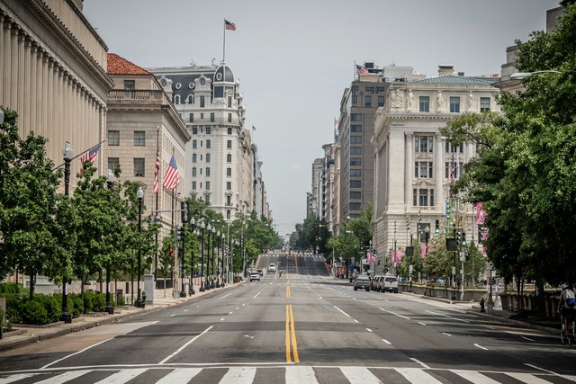 A view down an empty main street of Washington DC. The street is lined by trees, a few cars, many American flags, and white stone buildings, many with columns. Image at Offices.net.