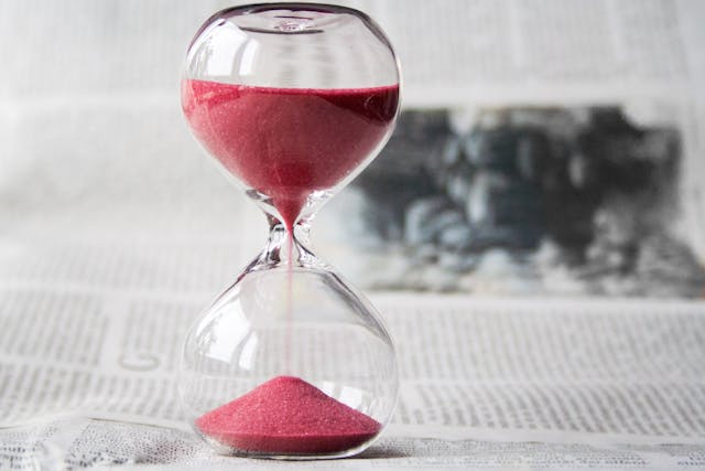 A close-up view of an hourglass with red sand flowing down. The hourglass is resting on a newspaper. Image at Offices.net.