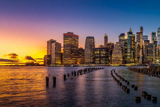 A shore-level view across the water of Hudson Bay during dusk towards the New York City skyline. Many of the city’s skyscrapers have their lights on and the whole scene is dramatically framed by and orange, pink, and purple sunset. Image at Offices.net.