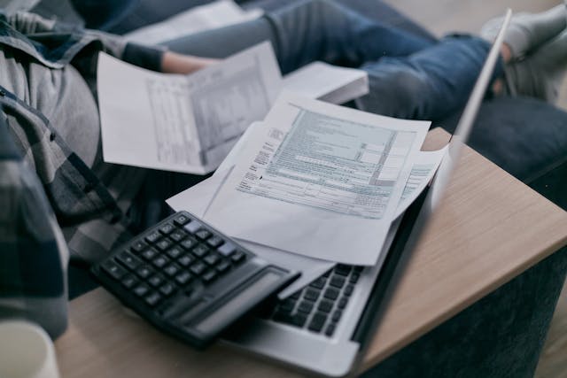 A businessperson in casual dress reclines on a couch with papers in their lap and reaches for a mug on a small desk affixed to the sofa’s right arm. The small desk also has a laptop on it with the screen angled towards the seated person, and there's a calculator and a couple of papers resting on the laptop, too. Image at Offices.net.