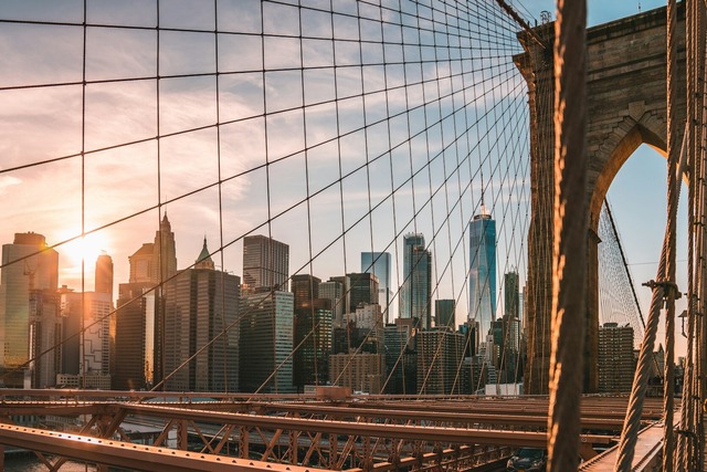 A late-day view from the Brooklyn Bridge walkway between safety wires towards the New York City skyline. One of the bridge’s tower supports is close by on the right of the view, and many tall skyscrapers clustered together dominate the left side of the view. The setting sun is bright shining through some white clouds and is just starting to go down below some of the buildings on the left. Image at Offices.net.