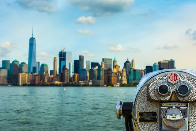 A daytime view from next to a coin-operated set of metal tourist binoculars, looking over the greenish blue Hudson River towards the skyline of Manhattan, New York City. Image at Offices.net.