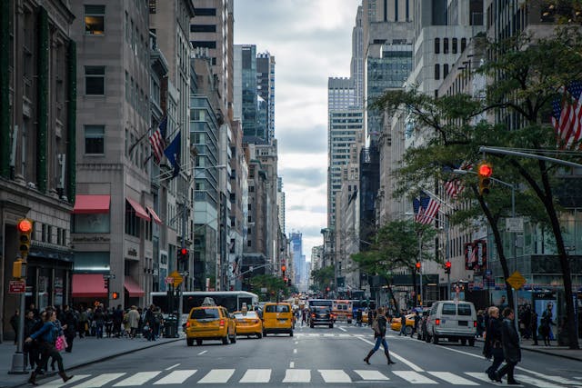 A daytime view down busy 5th Avenue in Midtown, Manhattan. Some people are walking across the street using white-striped crosswalks, and many are walking up and down the sidewalks on either side of the street. Tall buildings line the street and many cars including New York City’s iconic yellow cabs are driving down the street. Image at Offices.net.