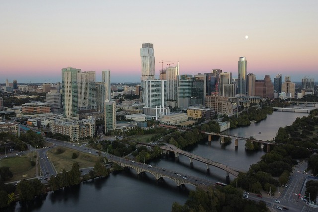 An aerial view across the city skyline of Austin, Texas at dawn. The Colorado River is winding alongside the city, and it is spanned by five visible bridges. The small white disc of the moon is just visible in the light peach colored sky, which gets darker towards the horizon. Image at Offices.net.