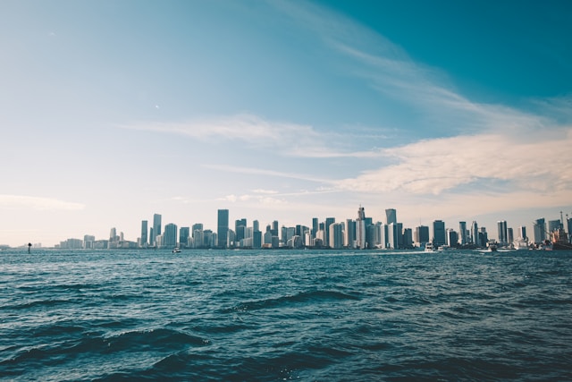 A daytime view from on the moderately calm water of the Biscayne Bay towards the distant Miami city skyline, which spans the width of the image. A few boats dot the water, and the sky is mostly light blue and sparsely streaked with white clouds. Image at Offices.net.