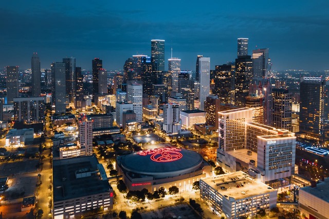 A nighttime aerial view of the city of Houston, Texas. Shorter buildings populate the near ground, and the taller skyscrapers line the background under a dark turquoise sky. Brightly lit roads criss-cross between buildings, many of which also have bright lights coming from their windows. Image at Offices.net.