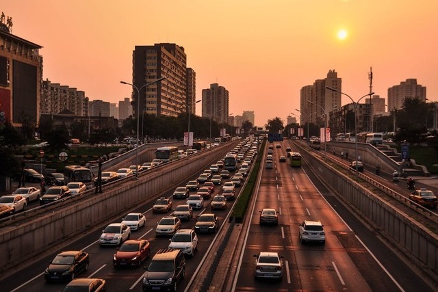 A view down the length of a stretch of freeway under an orange dusk sky with city skyscrapers in the distance. Cars are gridlocked coming towards the viewer on the left three lanes heading out of the city, and in the right three lanes, the traffic is more sparse heading towards the city. Image at Offices.net.