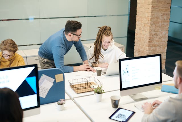 A male manager wearing a blue top and glasses leans on the desk of a smiling woman employee with dreadlocks and wearing a white hoodie. She has her laptop and a coffee cup in front of her, and her colleagues are working on their computers around her. Image at Offices.net.