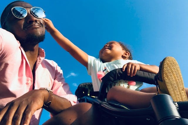 A smiling young father in a salmon colored collared shirt kneels next to his toddler’s pram as the child is laughing trying to take off his sunglasses, which shine with a mirror-reflective coating in the sun under a beautiful blue sky. Image at Offices.net.